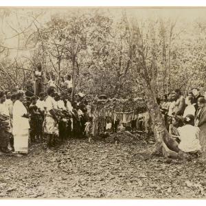 A group of people gathered around an earth grave. The grave has a floral cross and a feathered canopy.