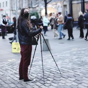 A woman wearing a face mask standing in front of a camera on a tripod, on the Royal Mile. 