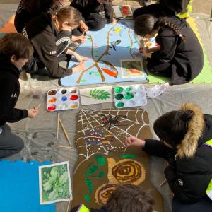 A group of children kneeling around wooden boards on the ground painting animals, insects and birds