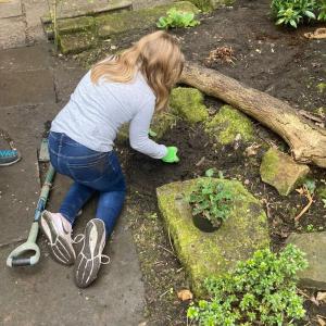 A girl from behind crouching on the ground wearing green garden gloves and preparing the soil for a new plant.