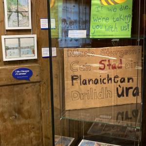 A tall museum case containing two climate action placards. Artworks by children are on the wall to the left of the case.