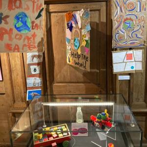 A museum case containing a glass bottle, plastic stickle bricks and a cart with wooden blocks. Cardboard climate action signs hang above the case