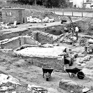 Excavation site in Cramond, Edinburgh. Two wheelbarrows in the foreground, a wooden shed in the background and 2 archaeologists at work beside a ladder.