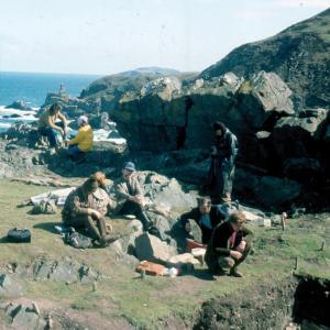 7 archaeologists sitting on large stones which was once an outer courtyard. The sea is in the background.