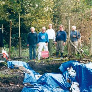 5 archaeologists pose for the camera in front of an archaeological site which is covered with blue tarpaulin